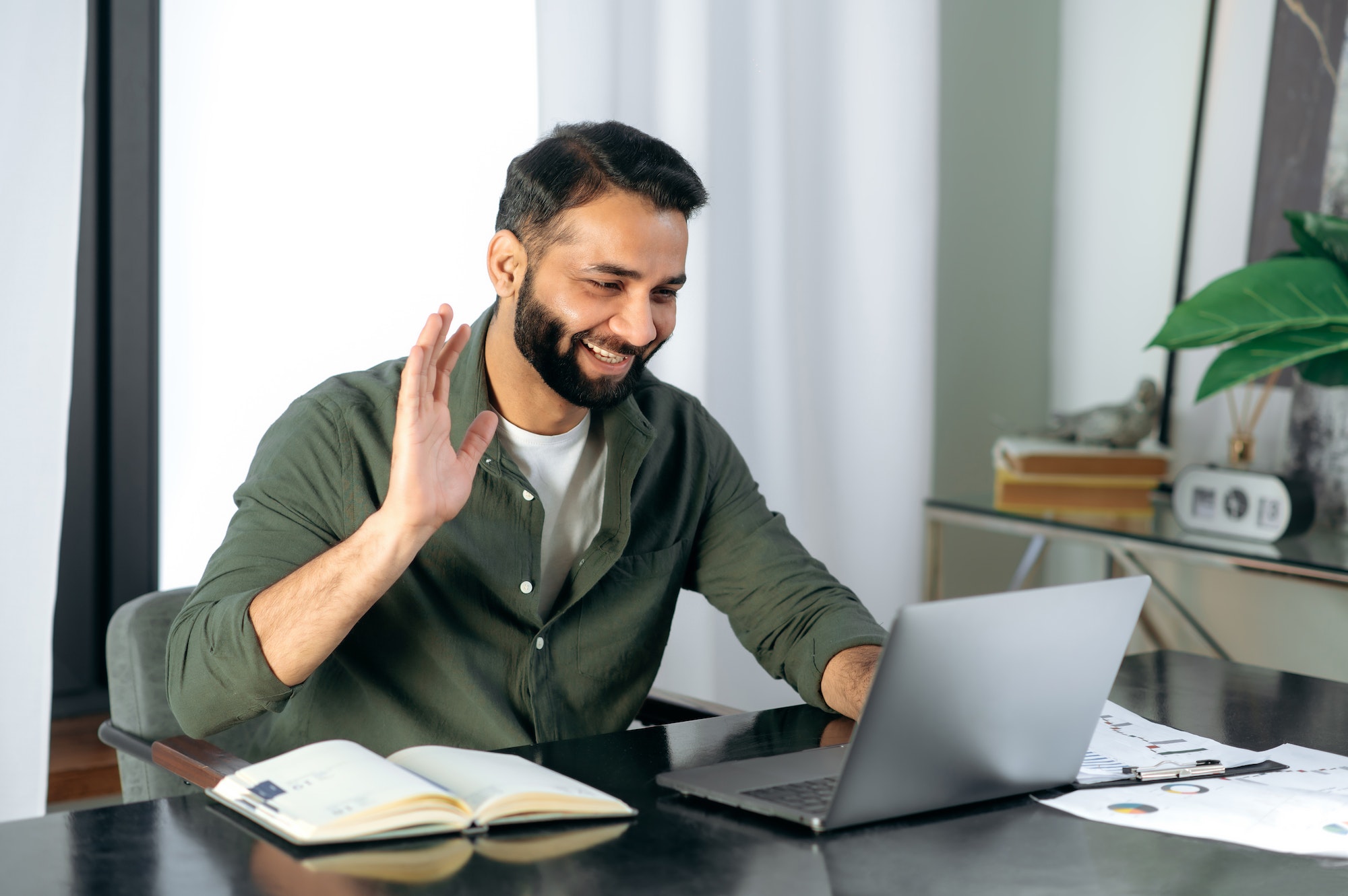 Man smiling and waving during a video call at a desk with an open notebook and documents nearby.
