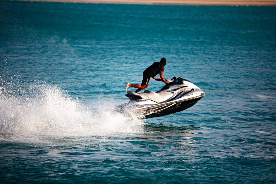 A person performs a jump on a jet ski, creating splashes in the vibrant blue water.