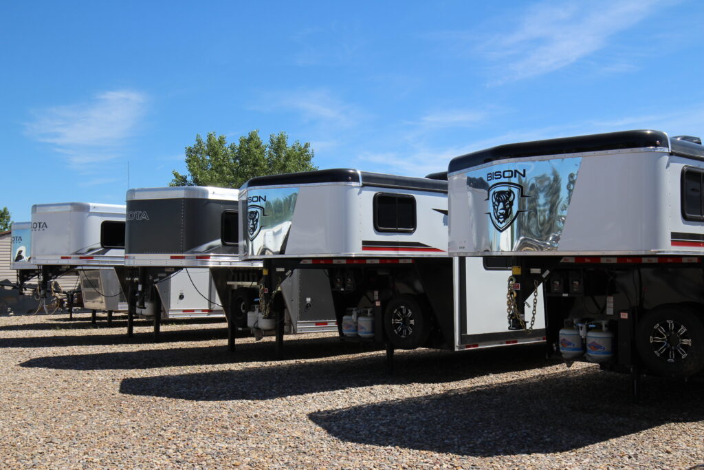 Row of horse trailers parked in a gravel lot under a clear blue sky, showcasing various brands and designs.