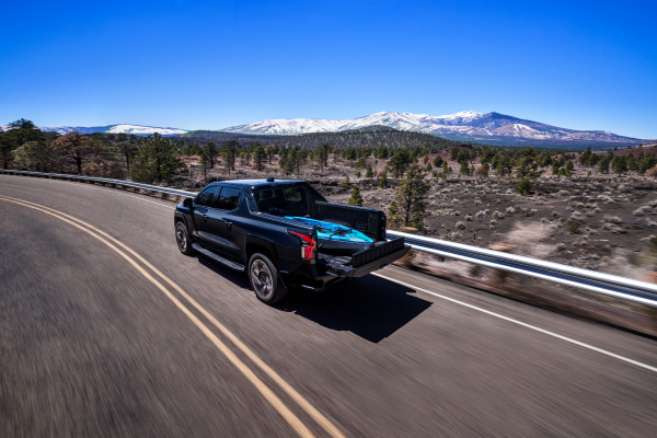 A black pickup truck drives along a winding road with snow-capped mountains and pine trees in the background.