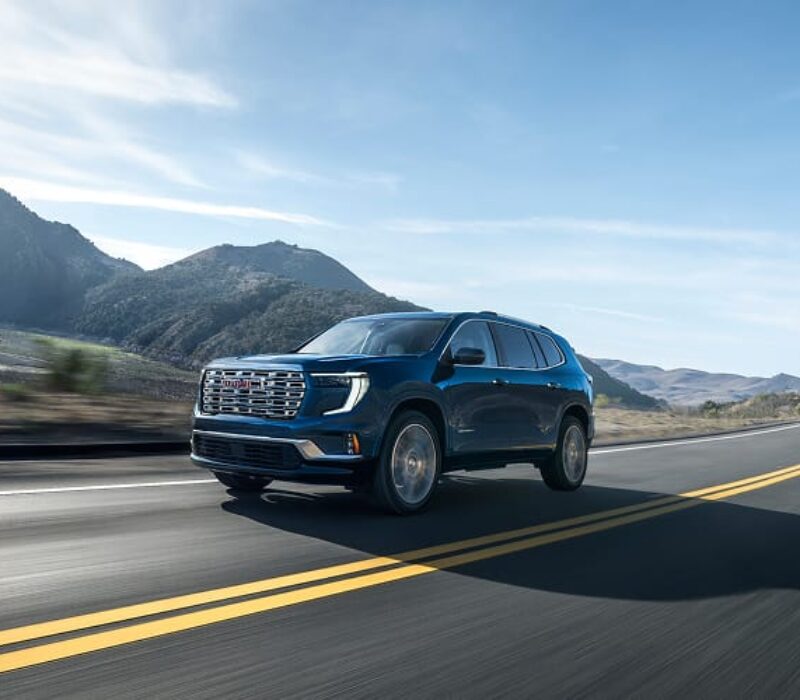 A sleek SUV drives along a winding road with mountains and a river in the background under a clear blue sky.
