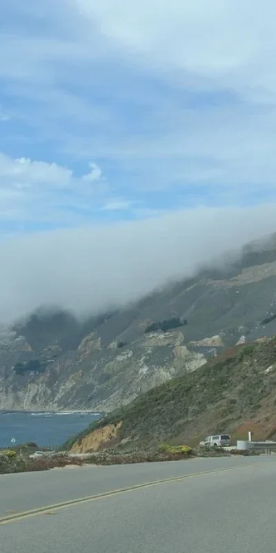 Coastal highway winding along a foggy cliffside with cars driving beside the ocean under a cloudy sky.